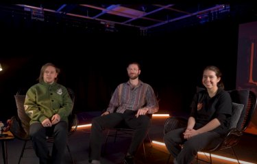 Photo of three people sitting on chair looking towards the camera. On the left, an anglepoise lamp sits on top of a side table. In the background there are neon strip lights.