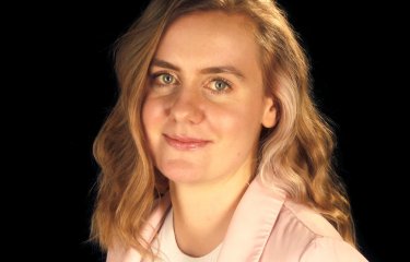 A portrait image of a woman, wearing a pink jacket and a patterned jumper