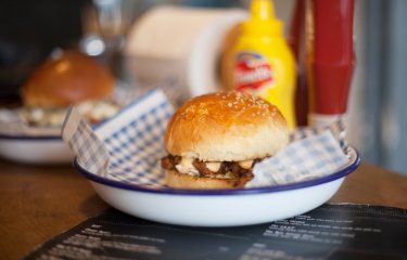 A burger in a white metal bowl with a bottle of mustard in the background