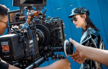 A man controlling a RED camera filming a women wearing sunglasses, a leather hat and jacket