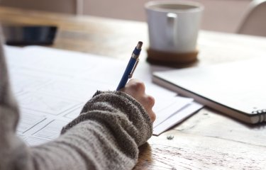 A hand holding a pen writing on paper with a mug in the background
