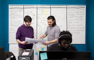 Two students stand in front of a whiteboard scrutinizing a piece of paper. A student sits in the foreground working on a laptop. 