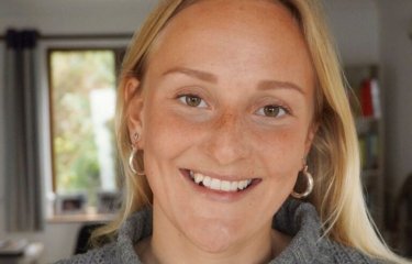 A blonde haired woman wearing a grey jumper smiles 
