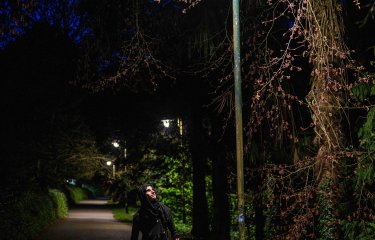A nighttime image of a person wearing a dark cloak in a road looking up at a streetlight