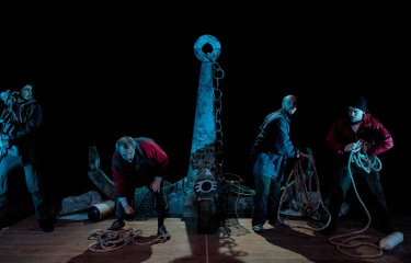 Five performers on stage under a very dim blue light with the large stage set of an anchor on stage in the middle of the group