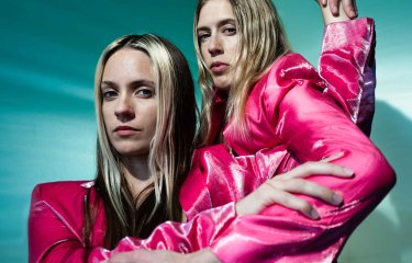 2 female performers wearing electric pink coats dancing against a teal green background