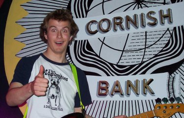 Image of a person holding a guitar in front of the colourful Cornish Bank logo painted on the wall