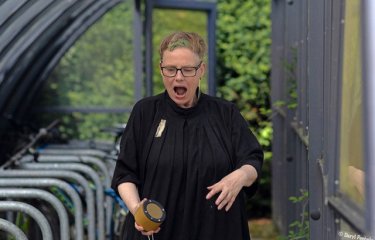 A performer located in a bike shed wearing black clothing holding a wireless speaker