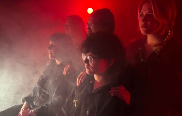 4 people seated and standing looking away from the camera on stage with a red light behind them with lots of smoke