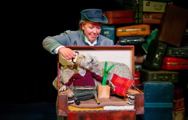 A person perfomring puppetry with a small puppet in an open suitcase with a background of books