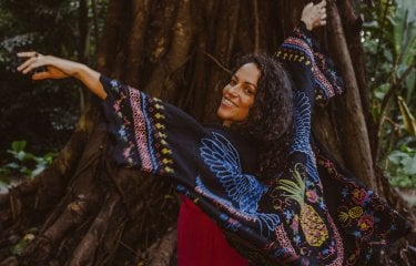 A performer in the woods in front od a tree with the arms in there air looking back over their shoulder towards the camera