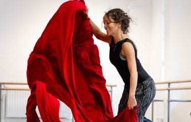 A dancer wearing dark clothing with a large piece of red fabric in a white dance studio