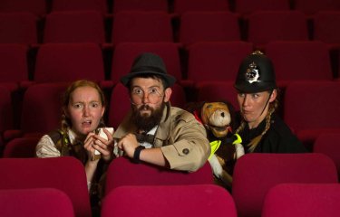 3 people sat on red cinema style chairs dressed up as police officers and 1 person has a puppet