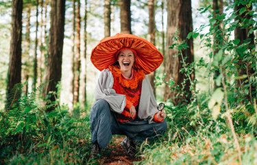 A person in the woods with a large orange fabric mushroom hat on