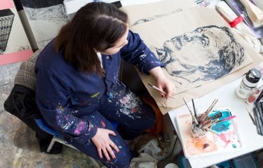 Photo of a young woman sat at a table with a drawing they are working on