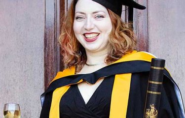 A graduate wearing her cap and gown, holding her degree certificate and a glass of wine
