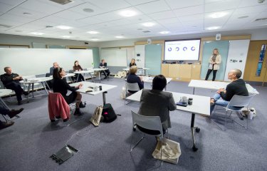 A student stands in front of a television screen and presents their business pitch to a panel of experts