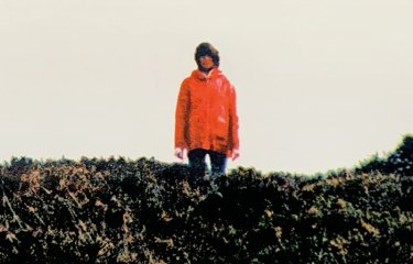 Film poster for ENYS MEN shows woman wearing jeans and red coat standing amongst yellow gorse. Text reads: From the director of BAIT, ENYS MEN, a film by Mark Jenkin