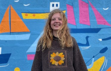 A girl wearing a brown jumper standing in front of a mural with boats