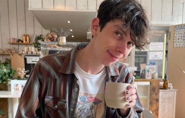 Photo of student Nathan Copeland wearing a graphic tee and checked shirt posing with a cup of coffee in a cafe.