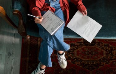 An aerial photo of a girl sat on a sofa with a notebook on her lap and a piece of paper next to her 