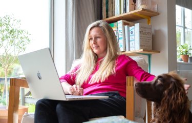 A female student in a pink top studying for her online university degree on her laptop in her sitting room while petting a dog