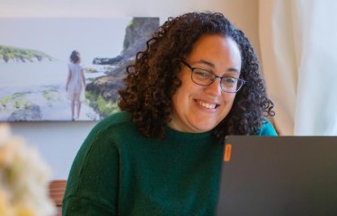 A woman with curly hair and glasses laughing at a laptop