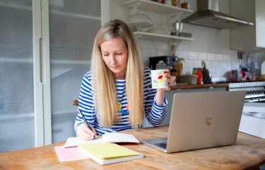 Woman at laptop writing script on paper, holding mug