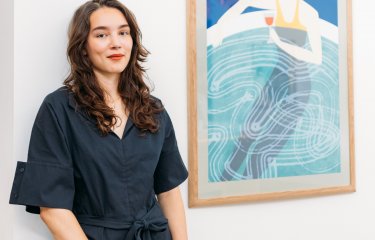 A young woman with dark hair standing next to a framed artwork of a woman drinking a glass of wine in a pool