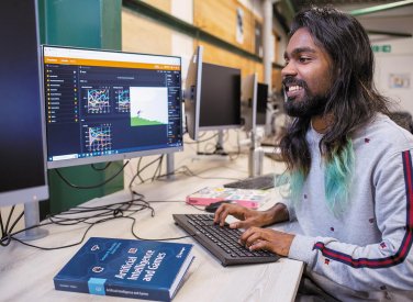 A man typing at a computer with an artificial intelligence for games book on the table