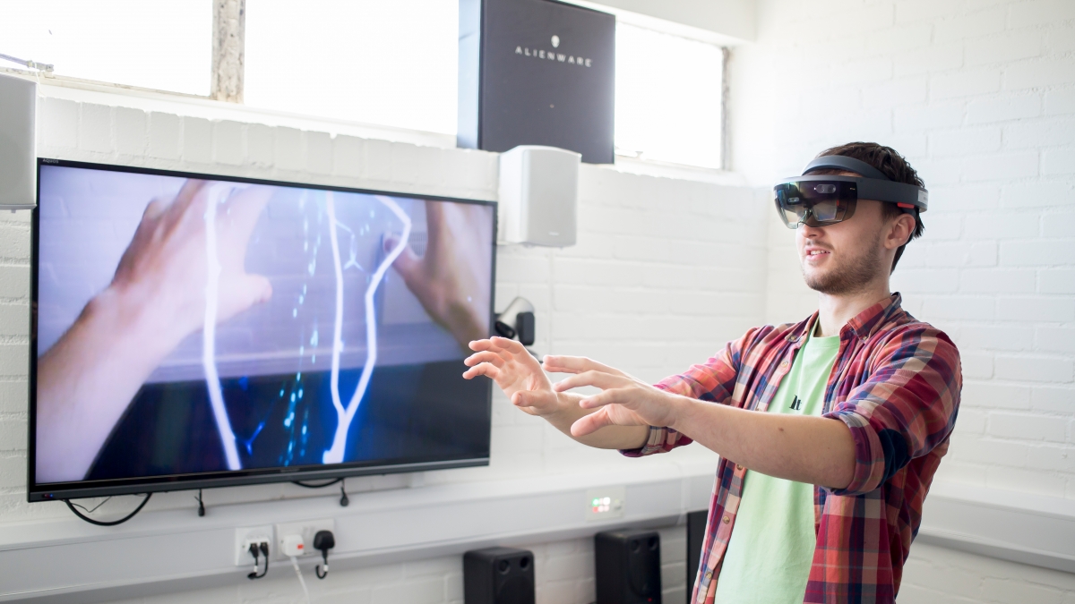 Student wearing virtual reality headset with arms extended in front and images of hands and electricity bolts on screen next to him.