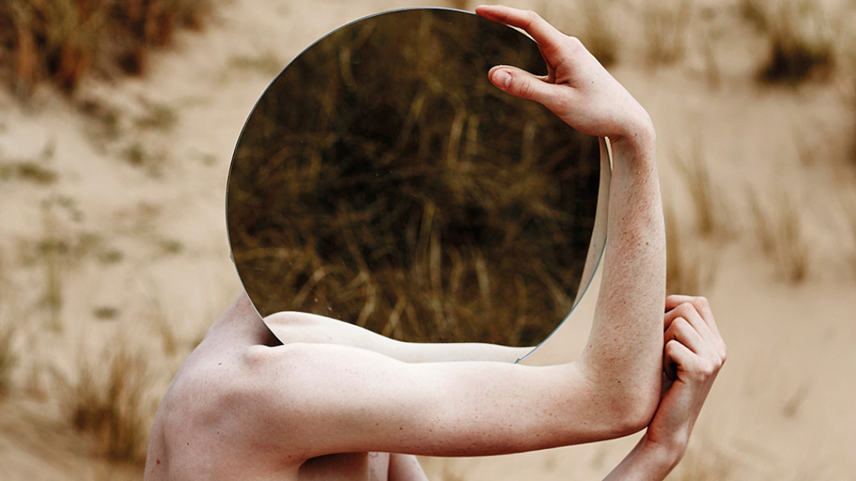 A topless man holding a mirror in front of his face