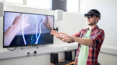 Student wearing virtual reality headset with arms extended in front and images of hands and electricity bolts on screen next to him.