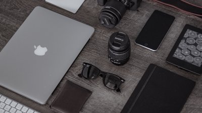 A desk with laptop, keyboard, camera, lens, smartphone.