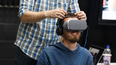 man seated wearing VR headset with another man stood behind him