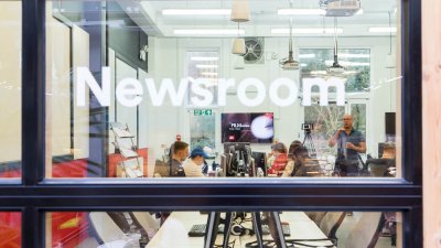 Through the window of the newsroom with lecturer talking to journalism students.