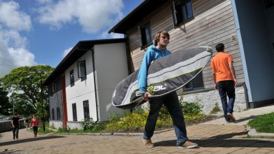 Student carrying surfboard outside student accommodation.