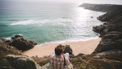 Man sat on the edge of cliff looking out to sun reflections on the water and coastline.