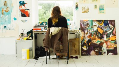 Back of student sitting at desk in front of window surrounded by artwork and paintings.