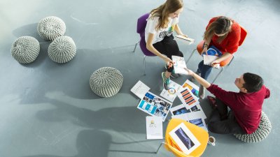 Falmouth University Business students sat on chairs on grey floor looking at work together.