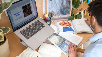 A man sat at a computer with fashion-related books and paper