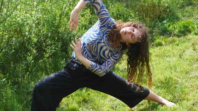 A person dancing outside on green grass with a bush in the background