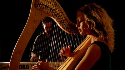 A close image of a harpist holding and playing the instrument with a person in the background playing an electric piano