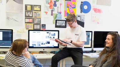 Three Falmouth University students sat round an iMac with graphic design posters on the wall