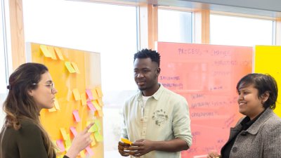 Three people standing in front of a yellow board covered in post-it notes