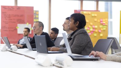 A group of people sat with laptops on a table, board of post-it notes behind them