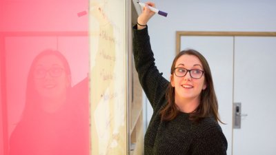 A woman wearing a black jumper, writing on a whiteboard with a pink panel