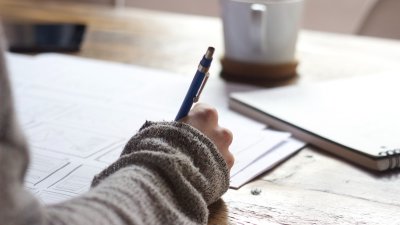 A hand holding a pen writing on paper with a mug in the background