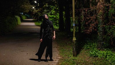 A nighttime image of a person wearing a dark cloak in a road looking up at a streetlight