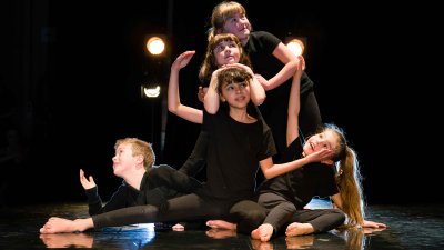 A group of mixed age young dancers posing on stage looking at the camera with some theatrical lighting in the background